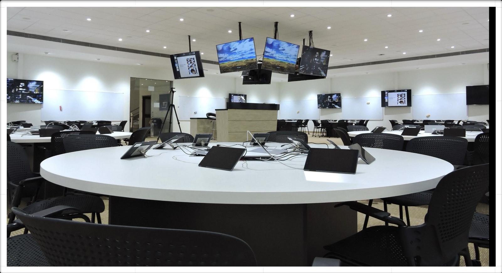 Studio AIR classroom: circular tables with tablets, an overhead cluster of 65-inch displays, writable walls, and a central instructor kiosk.